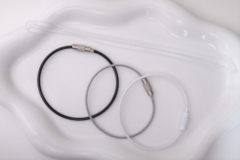 May include: Three circular cable key rings in black, silver, and white. Each ring features a thin, flexible metal wire construction with a threaded, cylindrical screw-lock closure mechanism made of metal for secure fastening of keys or tags.