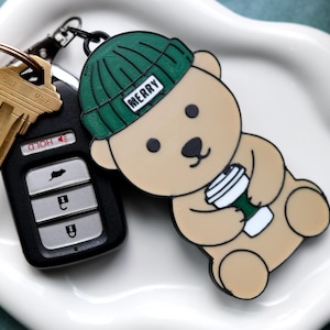 May include: A key set with a car key and a black key fob next to a cartoon bear keychain. The bear is wearing a green beanie with the word "MERRY" on it and holding a coffee cup. The keychain is tan and white.