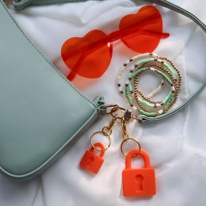 May include: A light green handbag with a shoulder strap, paired with orange heart-shaped sunglasses and a collection of beaded bracelets. Two orange padlocks hang from the bag's hardware, suggesting a fashion or accessory theme.