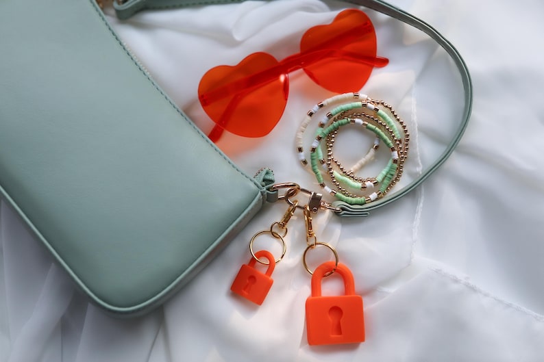 May include: A light green handbag with a shoulder strap, paired with orange heart-shaped sunglasses and a collection of beaded bracelets. Two orange padlocks hang from the bag's hardware, suggesting a fashion or accessory theme.