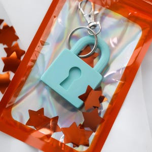 May include: A light blue padlock keychain with a silver clasp, packaged in a clear orange pouch. The keychain is surrounded by orange star-shaped confetti, creating a playful and decorative presentation. The background is white.