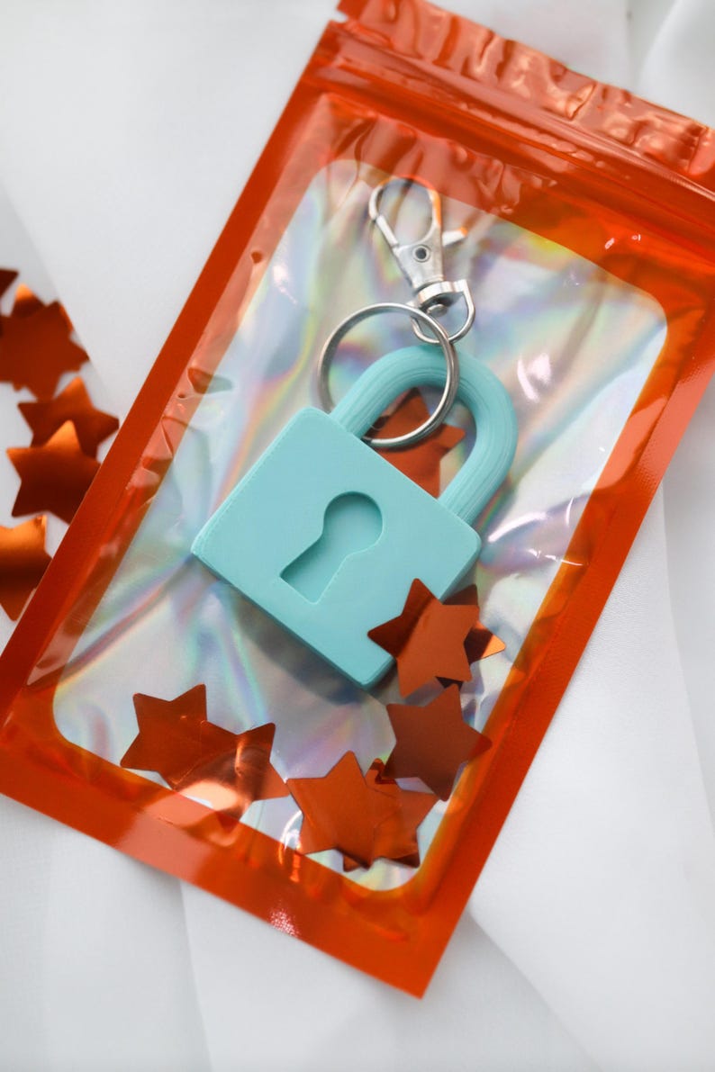 May include: A light blue padlock keychain with a silver clasp, packaged in a clear orange pouch. The keychain is surrounded by orange star-shaped confetti, creating a playful and decorative presentation. The background is white.