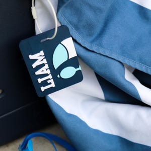 May include: A rectangular dark blue luggage tag with the name LIAM in white block letters and a light blue and white graphic of a swimming fin. The tag is attached by a white plastic loop to a blue and white striped fabric item.