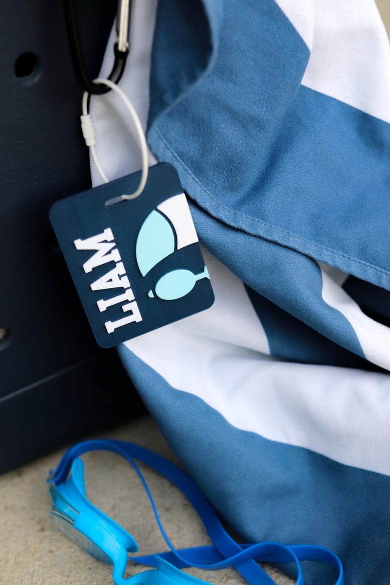 May include: A rectangular dark blue luggage tag with the name LIAM in white block letters and a light blue and white graphic of a swimming fin. The tag is attached by a white plastic loop to a blue and white striped fabric item.