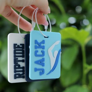 May include: Three rectangular luggage tags with white plastic loops. The front tag is light blue with the name JACK in dark blue letters and a white swimmer graphic. The middle tag is dark blue. The back tag is white with the word RIPTIDE in dark blue.