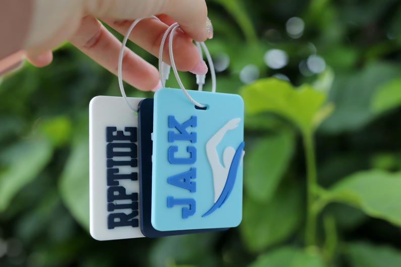 May include: Three rectangular luggage tags with white plastic loops. The front tag is light blue with the name JACK in dark blue letters and a white swimmer graphic. The middle tag is dark blue. The back tag is white with the word RIPTIDE in dark blue.