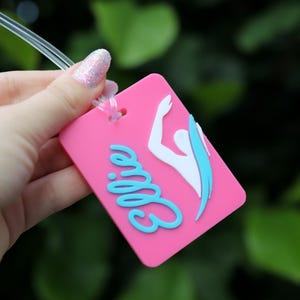 May include: A rectangular pink luggage tag featuring the name Ellie in raised light blue script lettering and a white silhouette of a swimmer with a light blue accent. The tag includes a clear plastic loop for attachment to personal items.