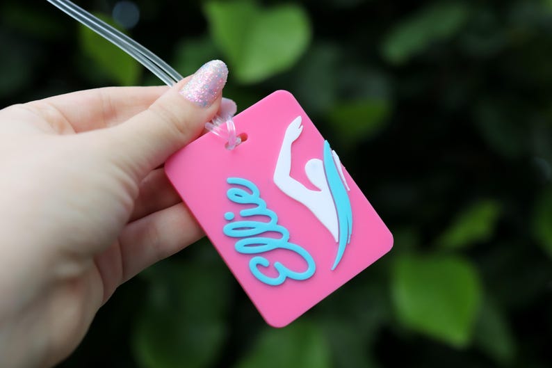 May include: A rectangular pink luggage tag featuring the name Ellie in raised light blue script lettering and a white silhouette of a swimmer with a light blue accent. The tag includes a clear plastic loop for attachment to personal items.