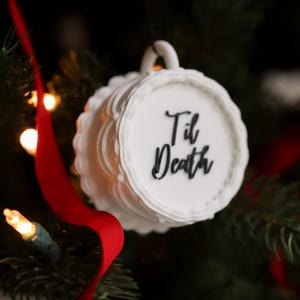 May include: White ceramic ornament shaped like a teacup, with the words "Til Death" in black script. The ornament has a scalloped edge and a handle. A red ribbon and Christmas tree lights are in the background.
