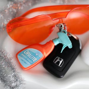 Könnte beinhalten: Eine Anordnung von Alltagsgegenständen auf einer weißen Oberfläche. Enthält orangefarbene herzförmige Sonnenbrillen, einen Schlüsselanhänger mit einer Miniatur-Sprühflasche, einen Autoschlüssel und ein weißes kabelloses Ohrhörer-Etui. Auf der Sprühflasche steht "OPALITE MAGICAL RESULTS".