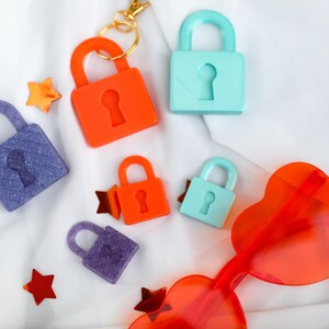 May include: An assortment of colorful padlocks in orange, blue, and purple, along with star-shaped confetti and heart-shaped sunglasses. The padlocks have keyhole details and are scattered on a white fabric background.
