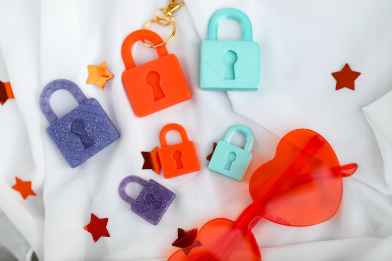 May include: An assortment of colorful padlocks in orange, blue, and purple, along with star-shaped confetti and heart-shaped sunglasses. The padlocks have keyhole details and are scattered on a white fabric background.