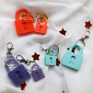 May include: Keychains in various colors, including orange, purple, and teal, shaped like padlocks. Each keychain has a keyhole design and is attached to a silver-colored clasp. The keychains are arranged on a white fabric with scattered star-shaped confetti.