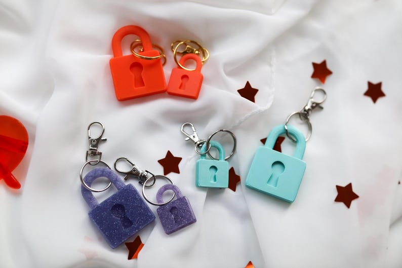 May include: Keychains in various colors, including orange, purple, and teal, shaped like padlocks. Each keychain has a keyhole design and is attached to a silver-colored clasp. The keychains are arranged on a white fabric with scattered star-shaped confetti.