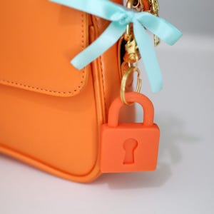 May include: Bright orange handbag with a matching orange padlock charm. The bag features a gold zipper and hardware, and a light blue ribbon bow. The padlock charm is attached to the bag with a gold keyring.