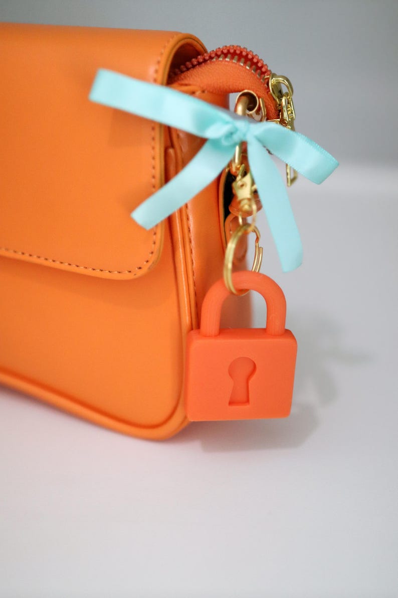 May include: Bright orange handbag with a matching orange padlock charm. The bag features a gold zipper and hardware, and a light blue ribbon bow. The padlock charm is attached to the bag with a gold keyring.