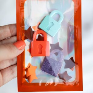May include: An orange cellophane bag containing three small, colorful padlocks in teal, orange, and purple. The purple padlock has glitter. The bag also contains orange star-shaped confetti. The bag is held by a person.