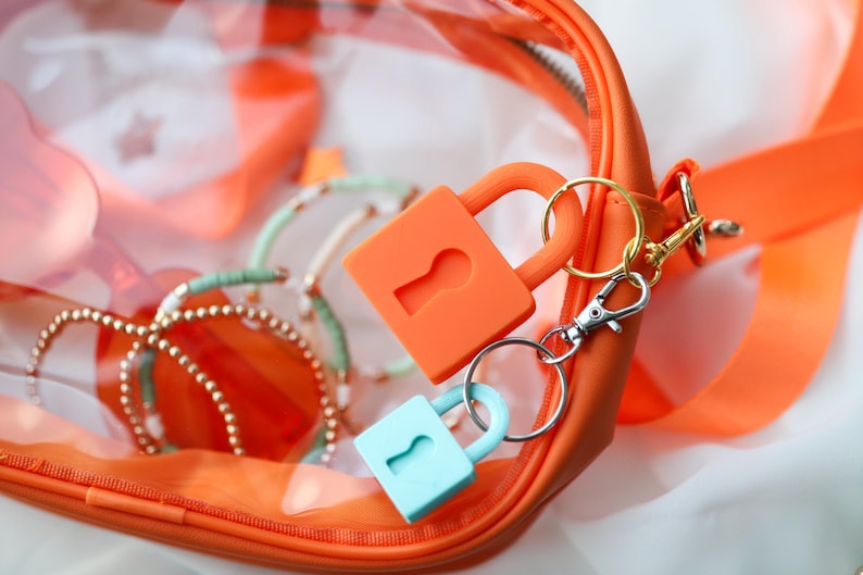 May include: An orange zippered pouch with a clear front, holding bracelets and two keychains. One keychain has an orange lock, the other a light blue lock. The keychains are attached to a gold and silver clasp.