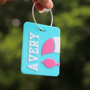 May include: A rectangular light blue luggage tag featuring the name AVERY in raised white letters. To the right of the name is a pink and white graphic of a sailboat. The tag is attached to a thin white circular ring for easy attachment.