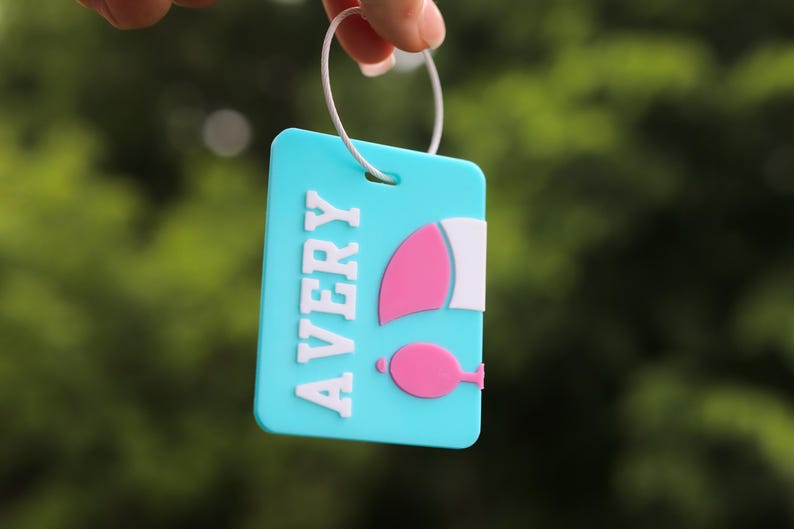 May include: A rectangular light blue luggage tag featuring the name AVERY in raised white letters. To the right of the name is a pink and white graphic of a sailboat. The tag is attached to a thin white circular ring for easy attachment.