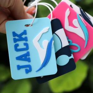 May include: A set of rectangular silicone luggage tags featuring raised designs of swimmers in various colors including light blue, navy, and pink. The light blue tag displays the name JACK in bold blue letters next to a white and blue swimmer graphic.