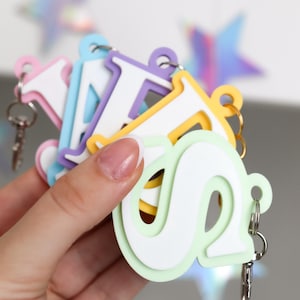 Pode incluir: Uma coleção de chaveiros de letras coloridas em tons pastel, incluindo rosa, azul, roxo, amarelo e verde. Cada chaveiro apresenta uma letra branca com contorno colorido e um chaveiro de metal. O fundo inclui formas de estrelas iridescentes.