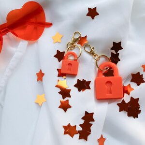 May include: Two orange padlock keychains with gold-colored hardware are displayed on a white fabric background, surrounded by star-shaped confetti in shades of orange and gold. A pair of orange heart-shaped sunglasses is visible.