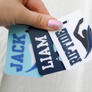May include: Three rectangular bag tags featuring raised lettering and graphics. One light blue tag displays the name JACK, a dark navy tag displays the name LIAM, and a white tag displays the name RIPTIDE with a black and navy swimmer silhouette.