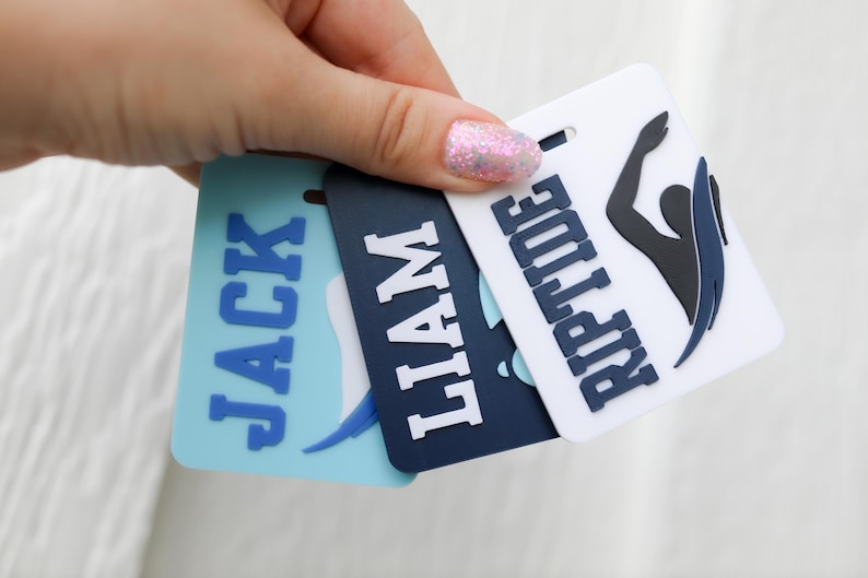 May include: Three rectangular bag tags featuring raised lettering and graphics. One light blue tag displays the name JACK, a dark navy tag displays the name LIAM, and a white tag displays the name RIPTIDE with a black and navy swimmer silhouette.