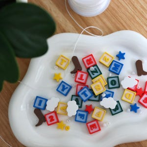 Op de afbeelding: Een verzameling kleurrijke vierkante letter- en cijferkralen, bruine cowboylaarsbedels, blauwe en gele sterbedels en witte wolkbedels verspreid over een witte, wolkvormige keramische schaal met een spoel wit draad in de buurt.