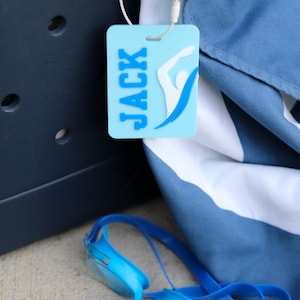 May include: A light blue rectangular luggage tag featuring the name JACK in dark blue letters and a white and blue swimmer graphic. The tag is attached with a white plastic loop to a dark blue and white striped fabric item near blue goggles.