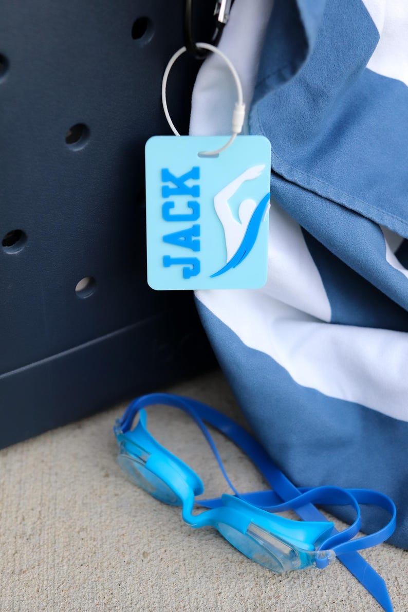 May include: A light blue rectangular luggage tag featuring the name JACK in dark blue letters and a white and blue swimmer graphic. The tag is attached with a white plastic loop to a dark blue and white striped fabric item near blue goggles.