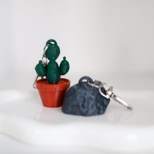 May include: Two keychains: a green cactus in a terracotta pot and a dark gray rock. The cactus keychain has a detailed design, while the rock keychain has a textured surface. Both have silver key rings.
