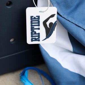 May include: A white rectangular bag tag featuring the word RIPTIDE in dark blue letters and a black graphic of a swimmer. The tag is attached with a white plastic loop to a blue and white fabric item, with blue swimming goggles visible below.