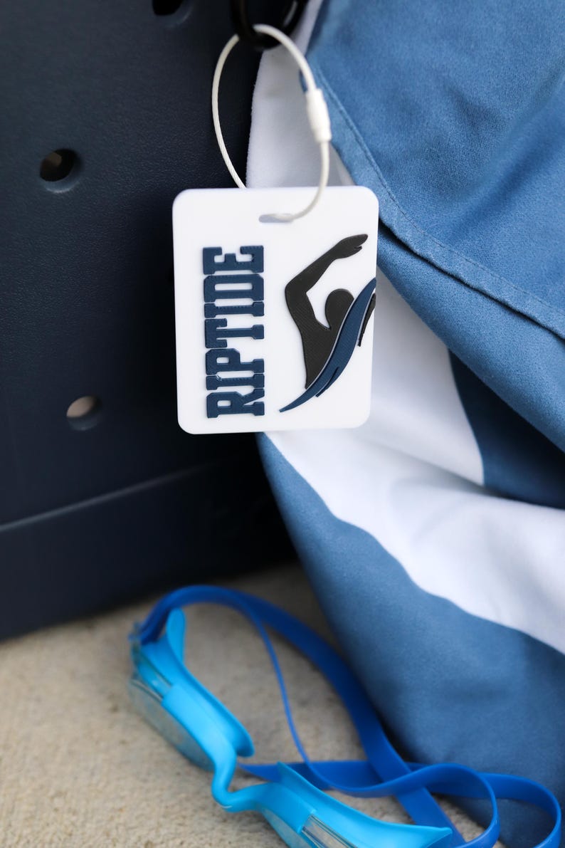 May include: A white rectangular bag tag featuring the word RIPTIDE in dark blue letters and a black graphic of a swimmer. The tag is attached with a white plastic loop to a blue and white fabric item, with blue swimming goggles visible below.