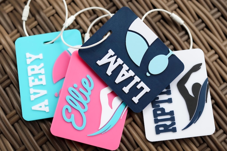 May include: Four rectangular luggage tags with white attachment loops. One is light blue with the name AVERY, one is pink with the name Ellie and a swimmer graphic, one is dark blue with the name LIAM and goggles, and one is white with RIPTIDE.