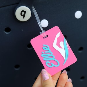 May include: A rectangular pink luggage tag featuring the name Ellie in light blue script lettering and a white silhouette of a swimmer with a light blue accent. The tag is attached to a dark navy blue perforated bag with a white circular button.