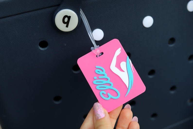 May include: A rectangular pink luggage tag featuring the name Ellie in light blue script lettering and a white silhouette of a swimmer with a light blue accent. The tag is attached to a dark navy blue perforated bag with a white circular button.