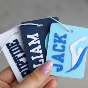 May include: Three rectangular luggage tags with rounded corners and white attachment strings. The tags are white, navy blue, and light blue, featuring raised block lettering that spells out RIPTIDE, LIAM, and JACK, with a swimmer graphic on the blue tag.