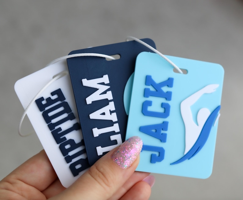 May include: Three rectangular luggage tags with rounded corners and white attachment strings. The tags are white, navy blue, and light blue, featuring raised block lettering that spells out RIPTIDE, LIAM, and JACK, with a swimmer graphic on the blue tag.