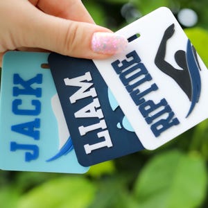 May include: Three rectangular plastic luggage tags in light blue, dark blue, and white. The tags feature raised lettering with the names Jack and Liam, and the word Riptide accompanied by a black and blue graphic of a swimmer in motion.