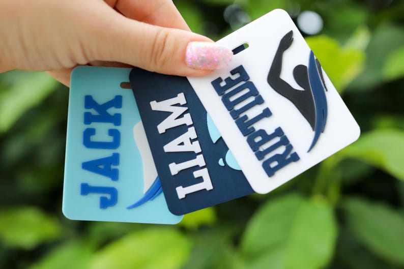 May include: Three rectangular plastic luggage tags in light blue, dark blue, and white. The tags feature raised lettering with the names Jack and Liam, and the word Riptide accompanied by a black and blue graphic of a swimmer in motion.