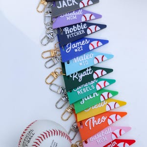 Könnte beinhalten: Eine Sammlung farbenfroher Baseball-Schlüsselanhänger mit Namen und Baseball-Grafiken. Jeder Schlüsselanhänger hat eine Wimpelform mit einem Baseball-Design. Ein Baseball ist ebenfalls im Bild enthalten.