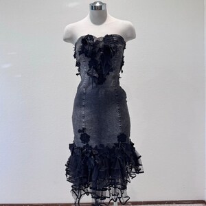 May include: A strapless denim dress with black floral and ruffle accents. The dress features a fitted bodice with a sweetheart neckline and a flared, ruffled hem. Black floral embellishments adorn the bodice and skirt, adding a touch of elegance to the garment.
