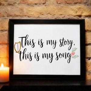 May include: A framed print with the words "This is my story, this is my song" in black script. A gold harp illustration and floral accents decorate the white background. A lit candle sits to the left.