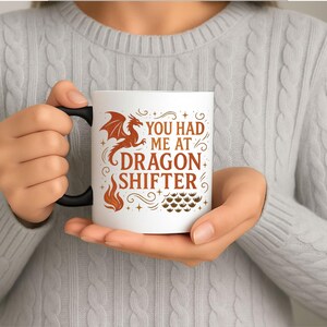 May include: White ceramic mug with a black handle featuring the text "YOU HAD ME AT DRAGON SHIFTER" in brown. A brown dragon graphic and flame design are also on the mug. The mug is held by a person.
