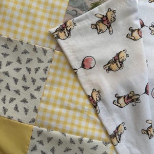May include: A patchwork quilt featuring alternating squares of yellow and white gingham, a cream fabric with a repeating grey bee pattern, and a white fabric printed with illustrations of Winnie the Pooh characters holding red balloons.