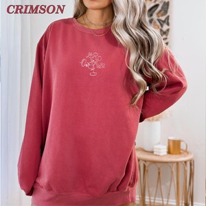Op de afbeelding: Karmizijnkleurige sweatshirt met een ronde hals en lange mouwen. De sweatshirt heeft een wit geborduurd Mickey Mouse-ontwerp op de voorkant. Het woord "CRIMSON" is in de linkerbovenhoek gedrukt.