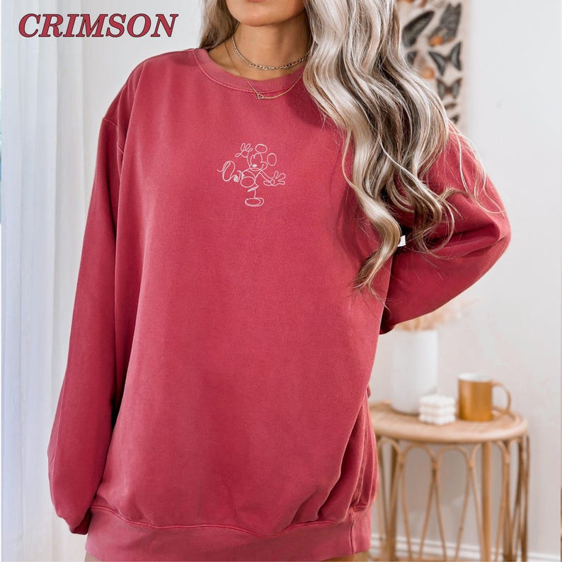 Op de afbeelding: Karmizijnkleurige sweatshirt met een ronde hals en lange mouwen. De sweatshirt heeft een wit geborduurd Mickey Mouse-ontwerp op de voorkant. Het woord "CRIMSON" is in de linkerbovenhoek gedrukt.