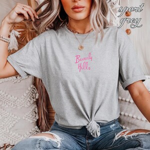 May include: A light gray t-shirt with the words "Beverly Hills" in pink script. The shirt is knotted at the waist and has rolled-up sleeves. The words "sport grey" are written in cursive in the upper right corner.